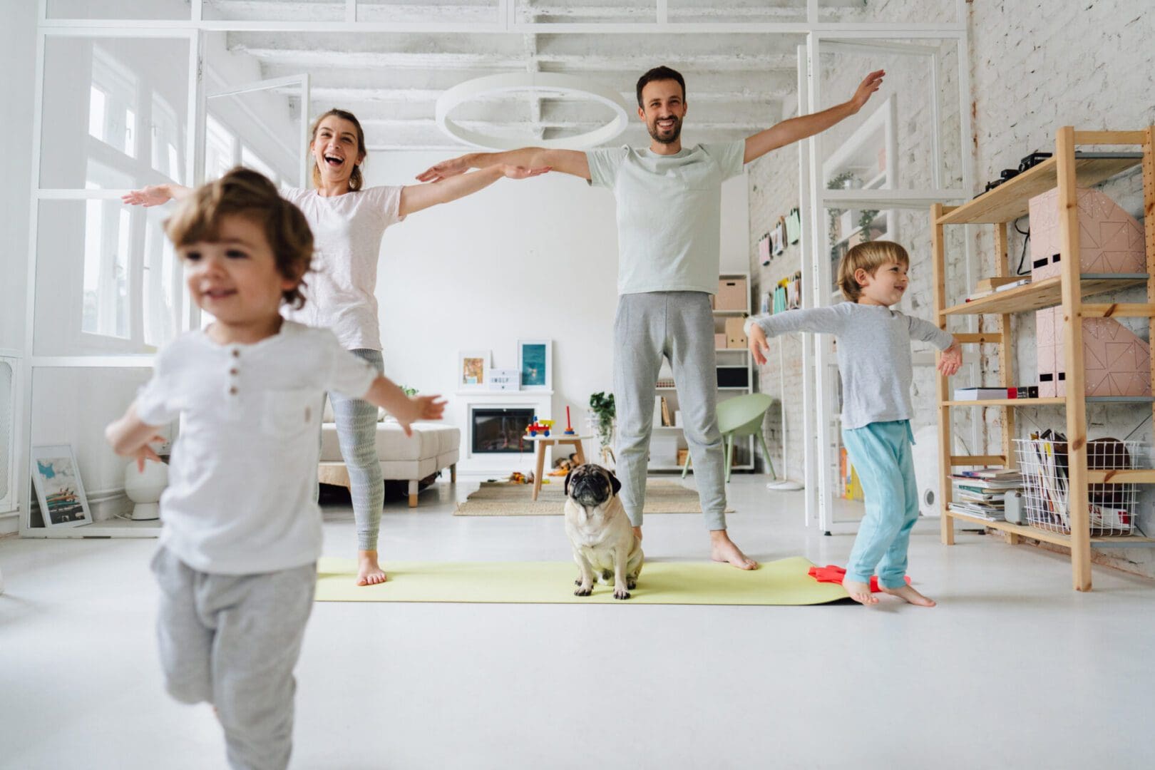 Photo of a young family exercising in the living room of their apartment; accompanied by their dog who is sitting on the yoga mat.